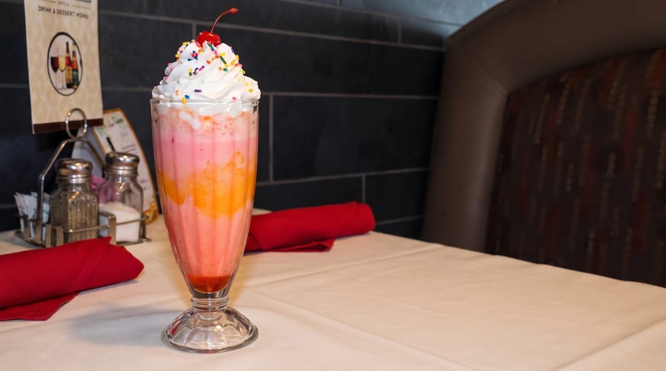 Pink and orange layered milkshake with whipped cream, sprinkles, and a cherry on a white tablecloth.