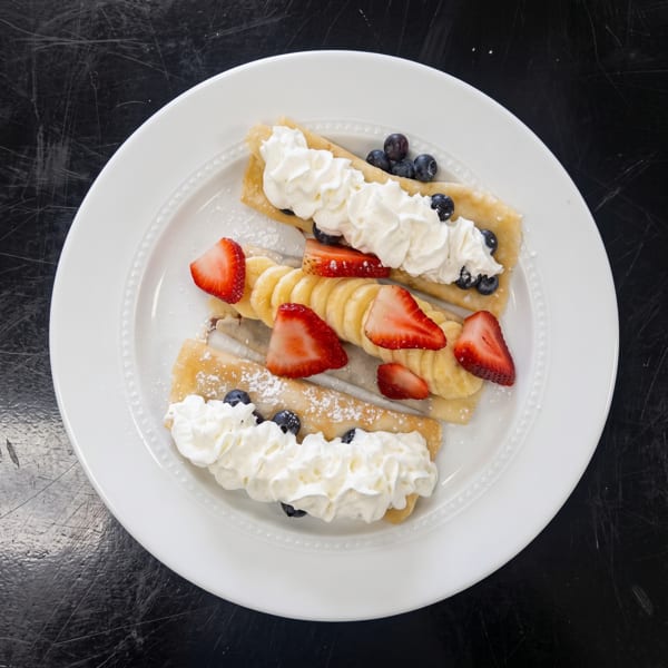 Three crepes topped with whipped cream, blueberries, sliced bananas, and strawberries on a white plate.