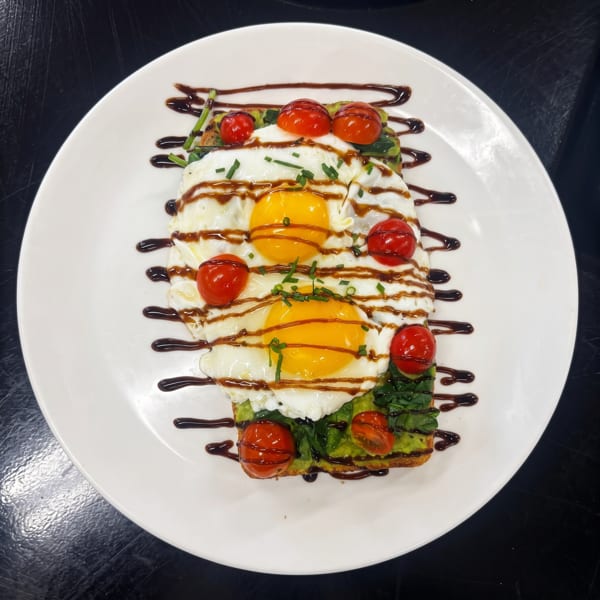 Avocado toast topped with two fried eggs, cherry tomatoes, chives, and balsamic glaze on a white plate.