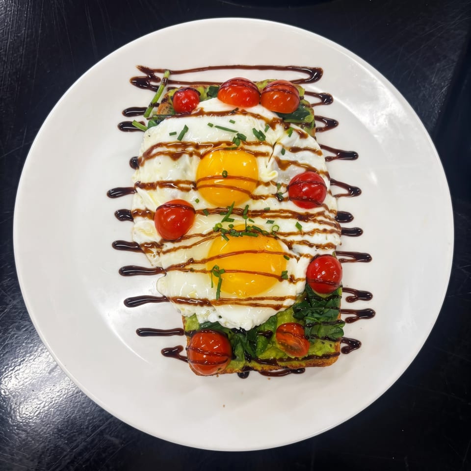 Avocado toast topped with two fried eggs, cherry tomatoes, chives, and balsamic glaze on a white plate.
