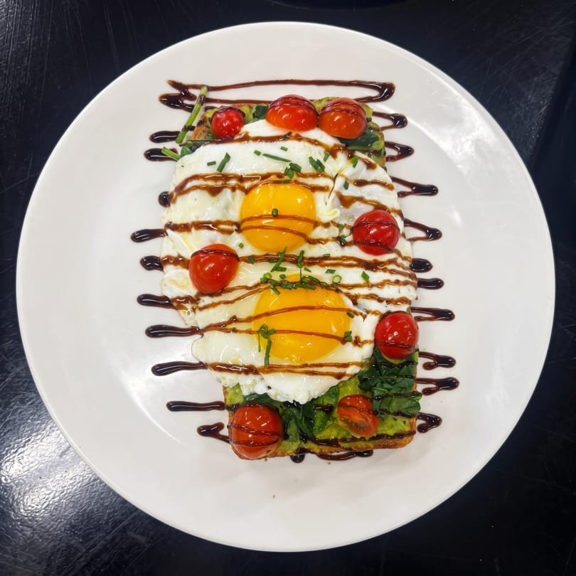 Avocado toast topped with two fried eggs, cherry tomatoes, chives, and balsamic glaze on a white plate.