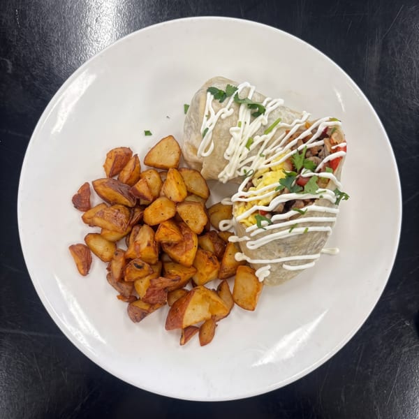 Breakfast burrito with scrambled eggs, meat, and vegetables, drizzled with white sauce, served with roasted potatoes.