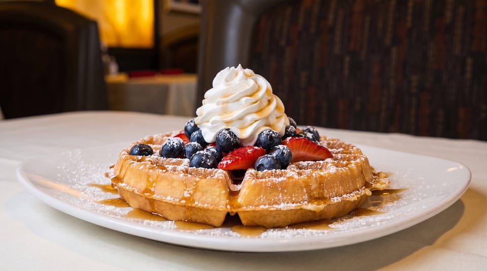 Golden waffle topped with whipped cream, blueberries, strawberries, powdered sugar, and maple syrup.