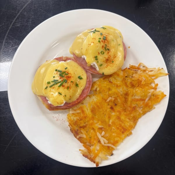 Two eggs Benedict with ham and hollandaise sauce, garnished with chives and paprika, served with hash browns.